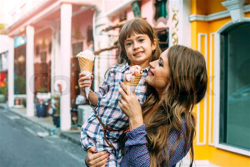 Mom with her 6 years old daughter walking along city street and eating ice cream in front of the outdoor cafe. Good relations of parent and child. Happy moments together, stock photo