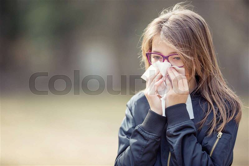Girl with allergy symptom blowing nose. Teen girl using a tissue in a park, stock photo
