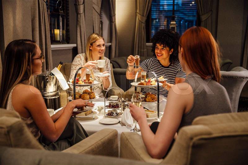 Four women are sitting together enjoying afternoon tea. They are talking and eating and they have champagne and tea. , stock photo