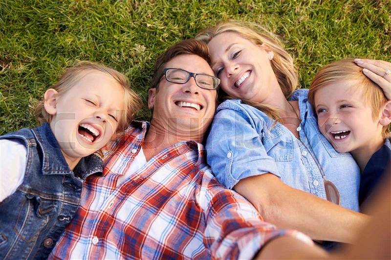 White family lies on grass taking selfie, camera out of shot, stock photo