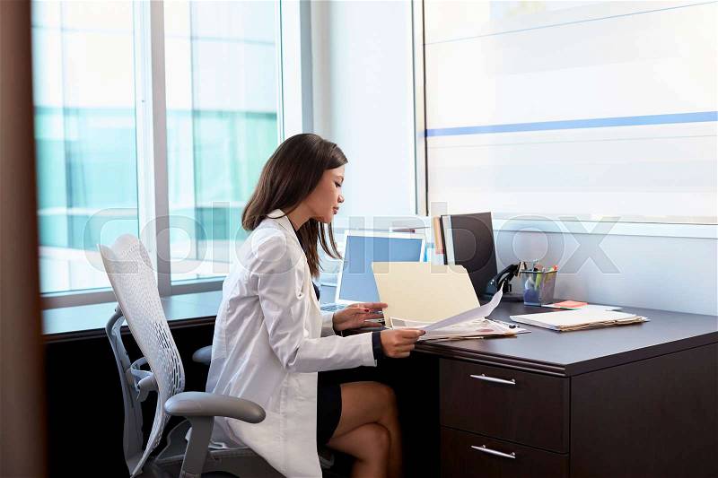 Female Doctor Wearing White Coat Reading Notes In Office, stock photo