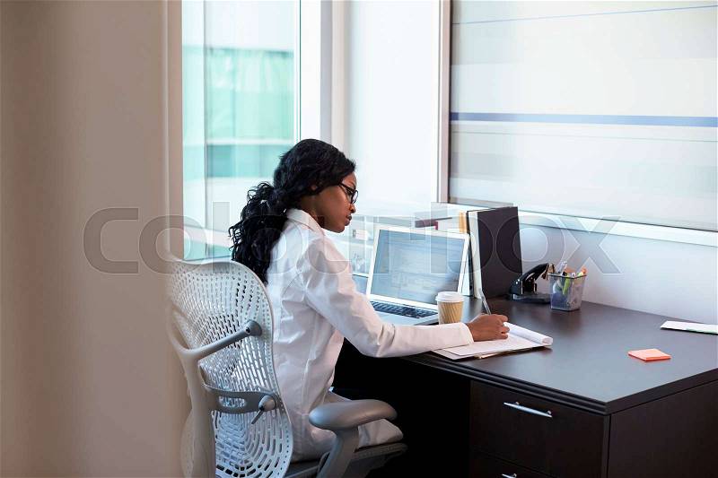 Female Doctor Wearing White Coat In Office Working At Desk, stock photo