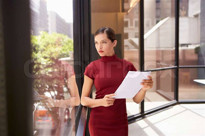 Businesswoman holding document, looking out of office window, stock photo