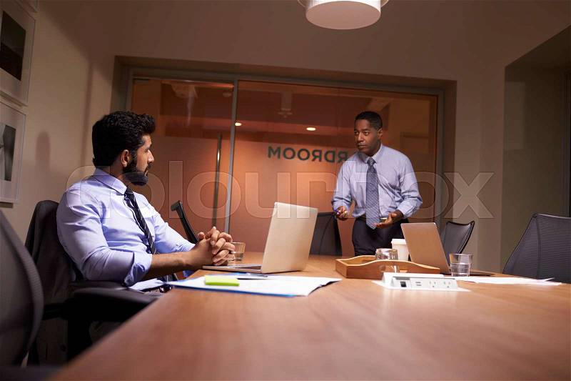 Businessman stands talking to work colleague late in office, stock photo