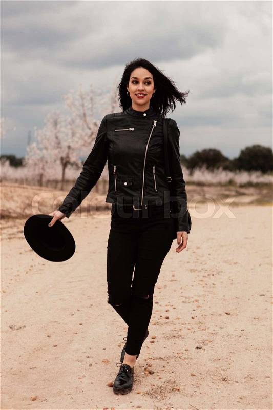Brunette woman with leather jacket walking on a path with flowered almond trees, stock photo