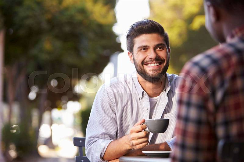 Two adult male friends sit talking over coffee outside cafe, stock photo