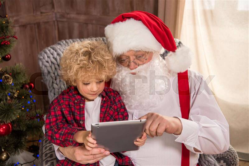 Santa Claus sitting on armchair with kid on knee and using digital tablet, stock photo
