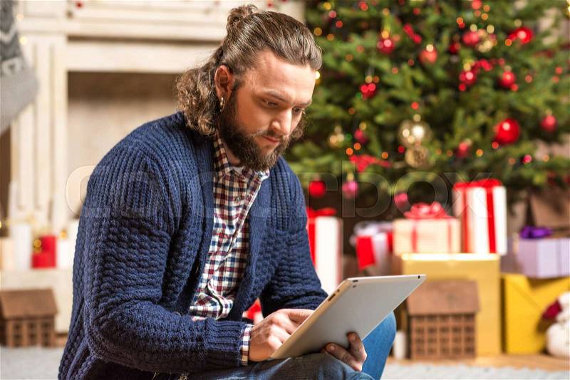 Young thoughtful man sitting on floor and using digital tablet in cozy room decorated for Christmas, stock photo
