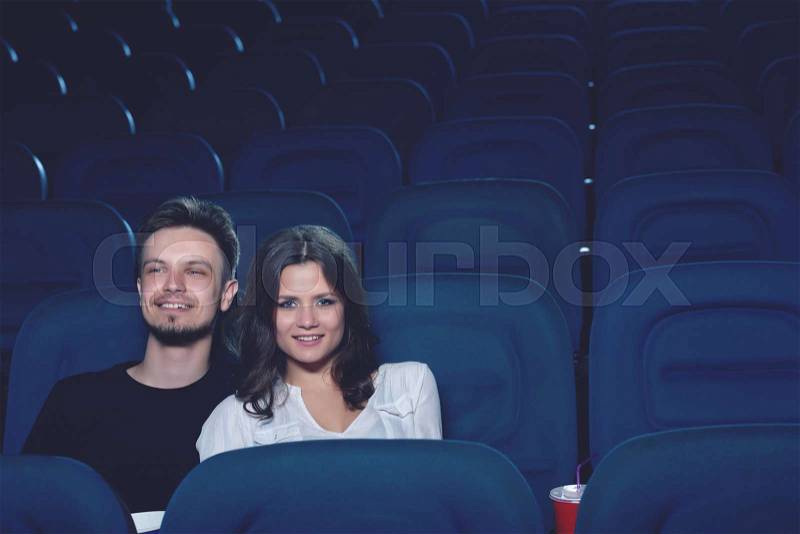 Front view of positive couple watching movie in cinema and looking at projector. Boyfriend embracing brunette girlfriend, sitting alone in big hall, stock photo
