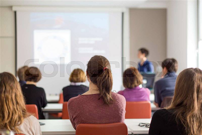 Female speaker giving presentation in lecture hall at university workshop. Rear view of unrecognized participants listening to lecture and making notes. Scientific conference event, stock photo