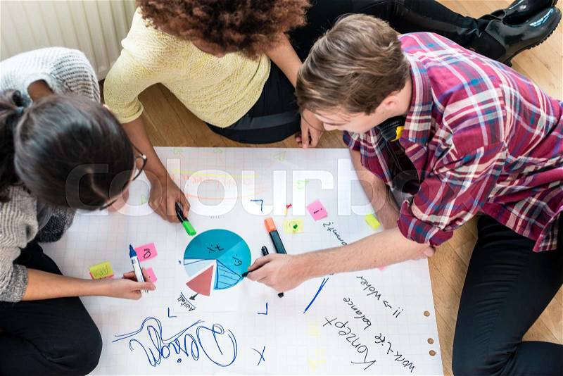 Three young people analyzing pie chart and writing observations on a large paper sheet during brainstorming session, stock photo