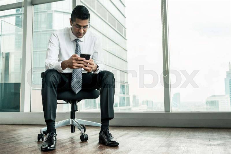 Indian businessman using a mobile phone while sitting down in a modern office, stock photo