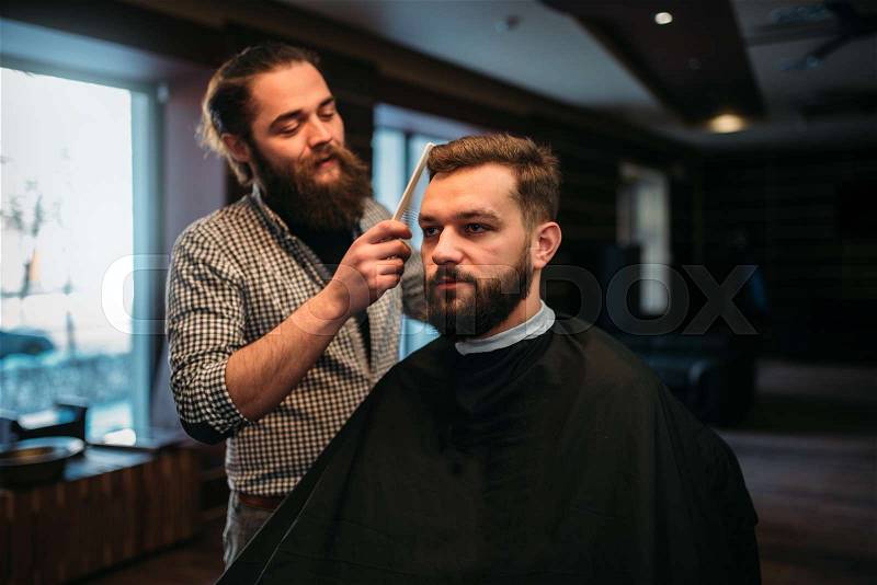 Barber combing hair of the client man in salon cape. Barbershop concept, stock photo