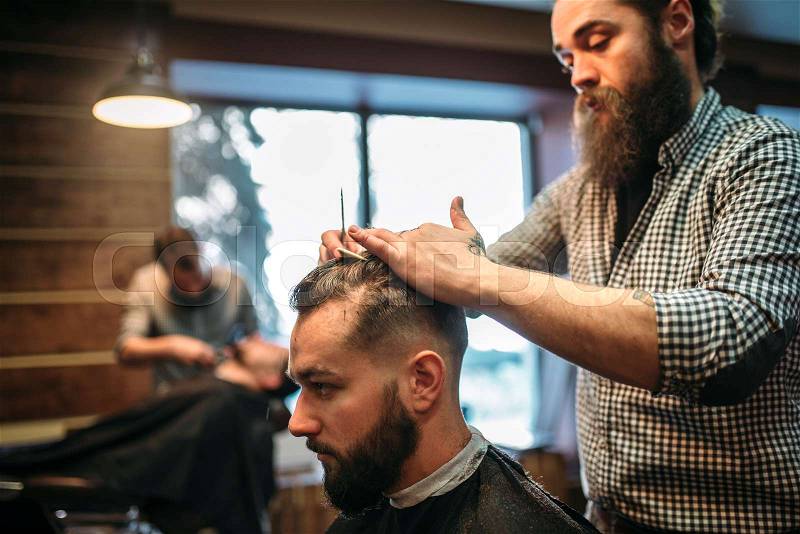 Bearded hairdresser making male hipster hairstyle. Client man in black salon cape visit barbershop, stock photo