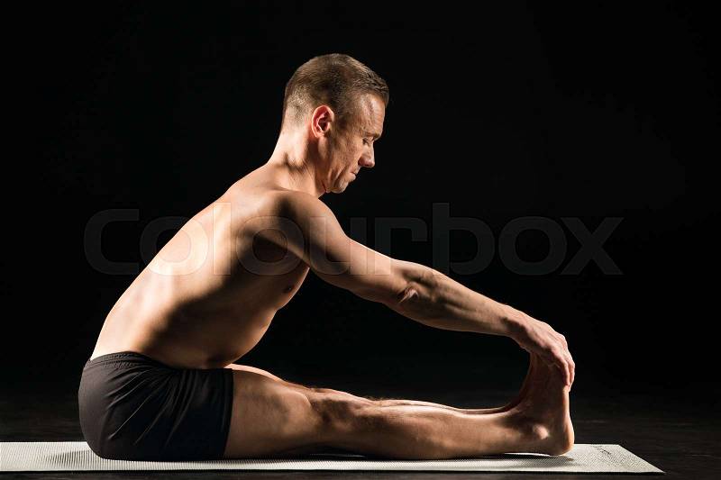 Man practicing yoga while sitting with straight legs and holding toes with hands, stock photo