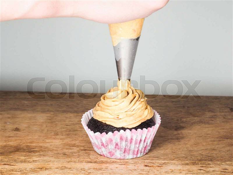 Professional cake baker applying frosting onto a chocolate cupcake on a wooden table, stock photo