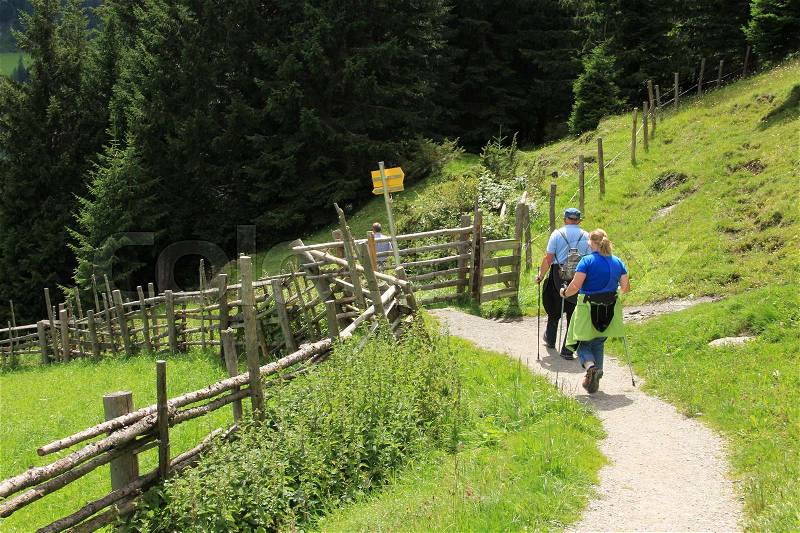 The couple, man and wife with walking sticks comes closer to a yellow signpost in the mountains in Austria in the beautiful summer, stock photo