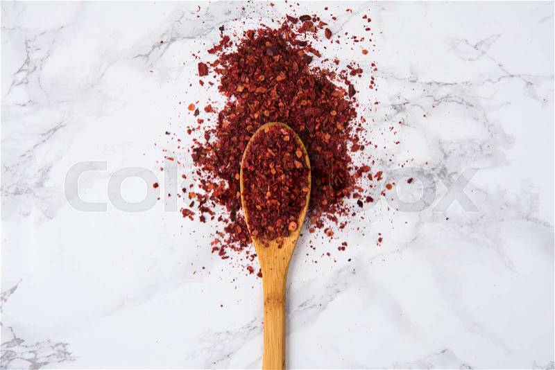 Red hot chilli pepper flakes in wooden spoon isolated on white marble background, stock photo