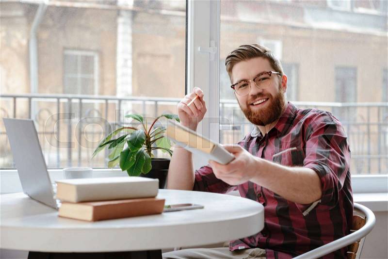 Image of happy bearded young man student sitting in cafe showing book to camera while using laptop computer. Looking at camera, stock photo