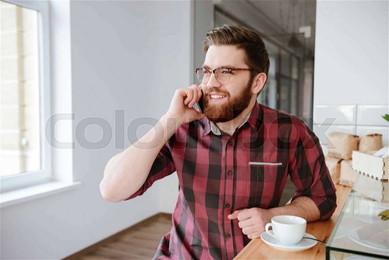 Young bearded man drinking coffee and talking on the phone while sitting at cafe, stock photo