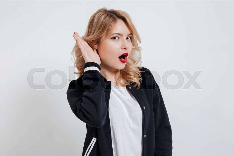 Portrait of a young cute girl trying to hear rumors holding hand at her ear isolated on the white background, stock photo