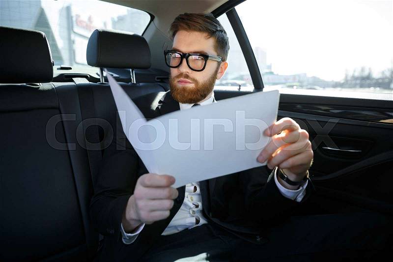 Serious businessman with documents driving on car back seat, stock photo