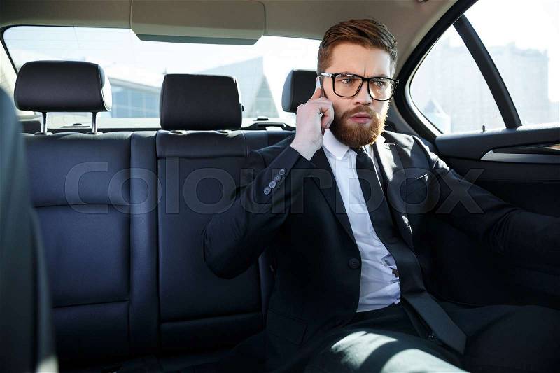 Portrait of an angry frustrated business man in eyeglasses talking on mobile phone while sitting in the back seat of car, stock photo