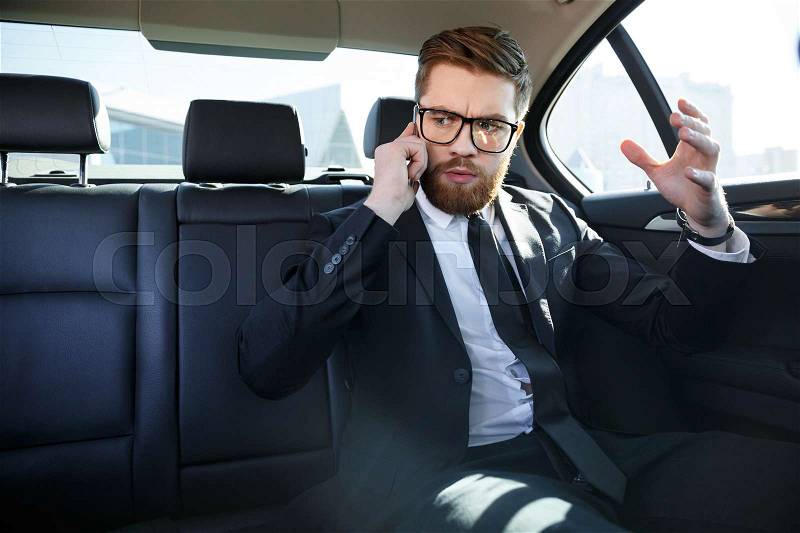 Disappointed bearded business man in eyeglasses talking on mobile phone in a back seat of a car, stock photo
