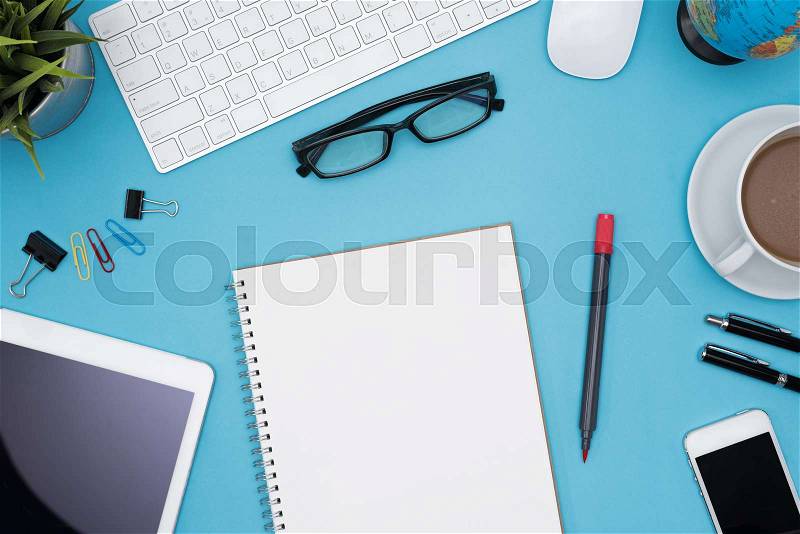 Modern blue office desk table with computer, tablet, cellphone, other supply, eye glasses, cup of coffee and blank notebook page for input the text. Top view, flat lay, stock photo