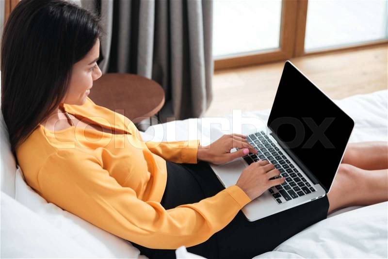 Side view portrait of a smiling businesswoman with laptop computer typing in bed at hotel, stock photo