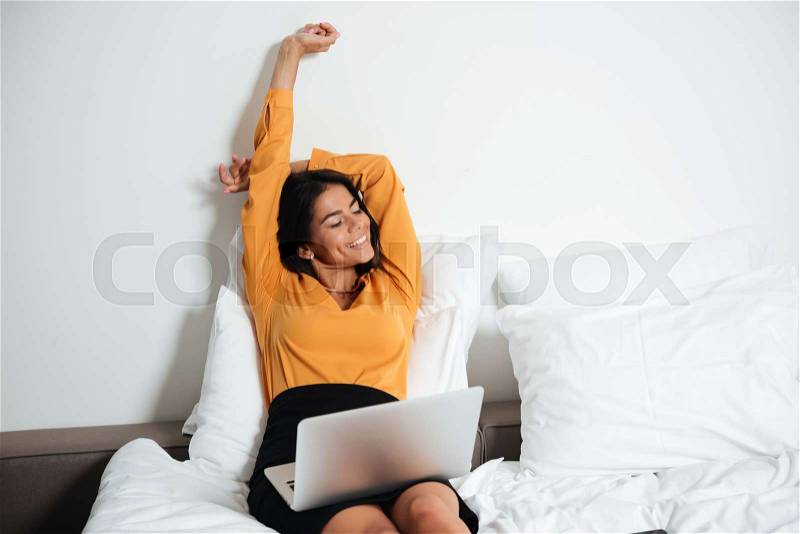 Happy young businesswoman laying in bed with laptop and stretching hands, stock photo
