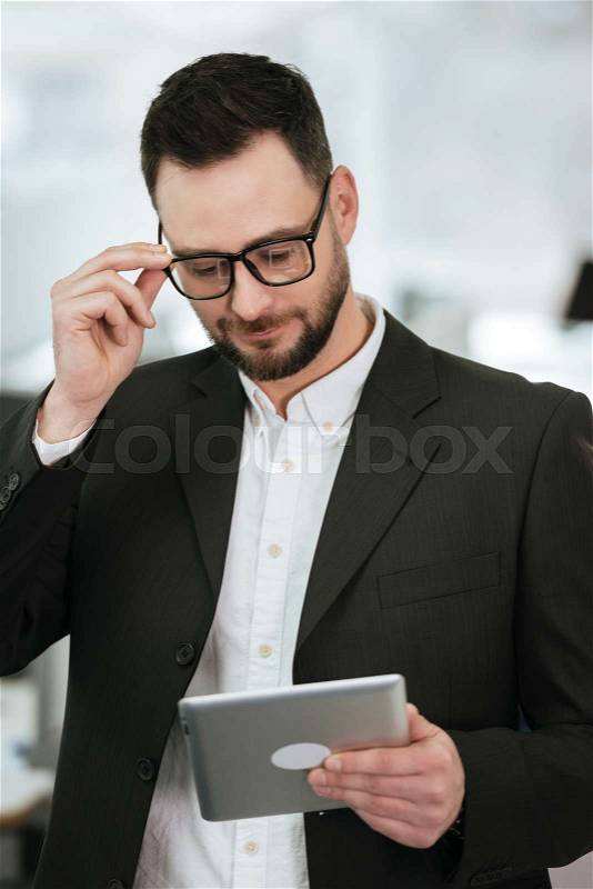 Vertical image of Pensive Bearded business man in suit and eyeglasses which using tablet computer in office, stock photo