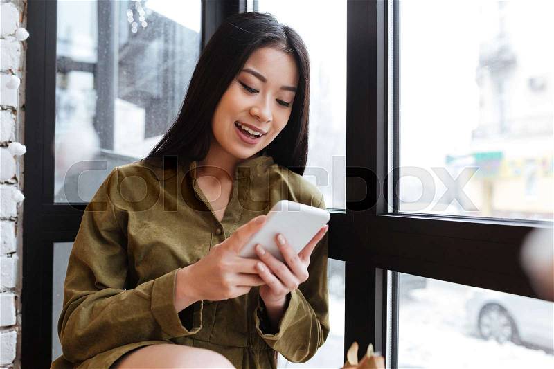 Picture of a beautiful asian young lady sitting at the cafe near window while chatting by her mobile phone. Looking aside, stock photo