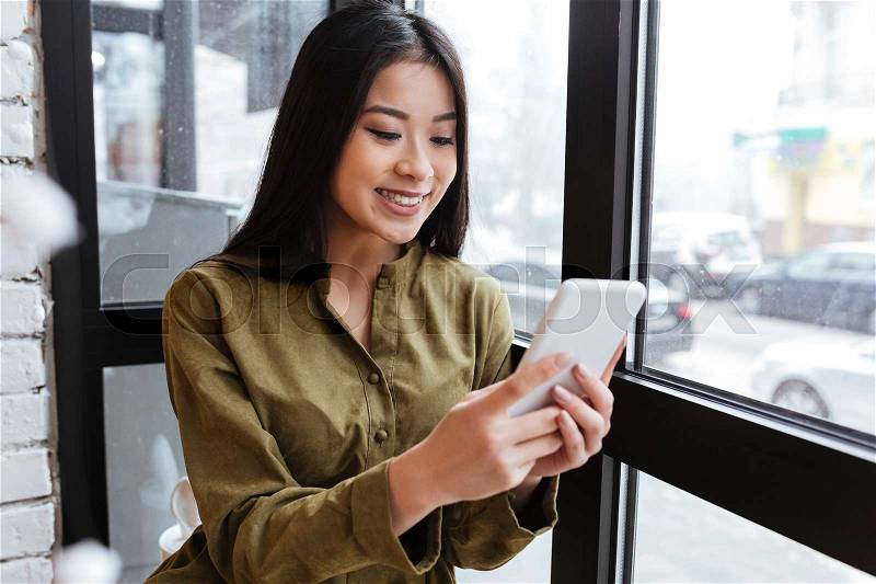 Image of a pretty asian young lady sitting at the cafe near window while chatting by her mobile phone. Looking aside, stock photo
