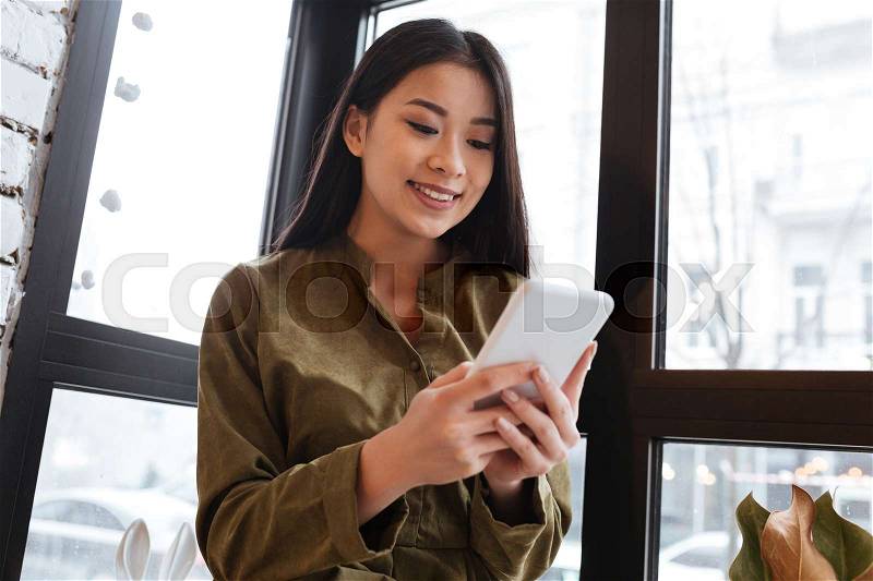 Image of an amazing asian young woman sitting at the cafe near window while chatting by her mobile phone. Looking aside, stock photo