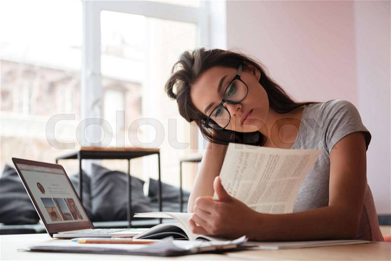 Photo of young amazing caucasian woman sitting indoors using laptop computer and reading magazine. Coworking concept, stock photo