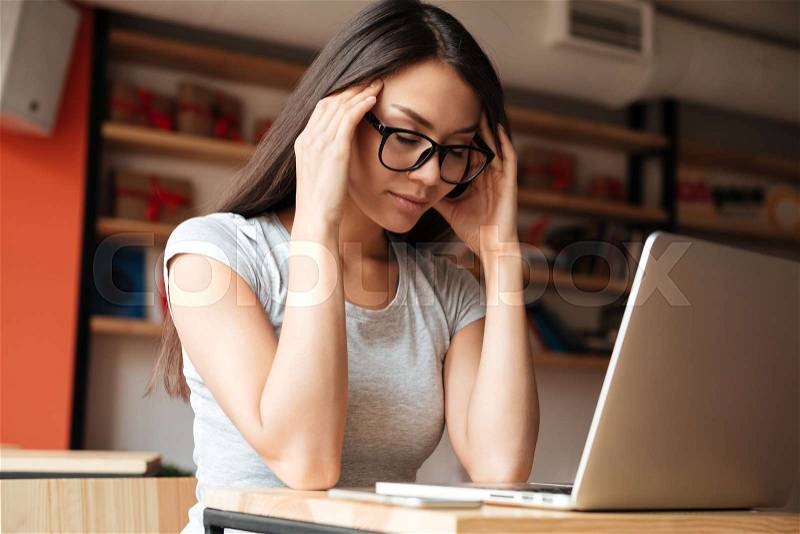 Photo of young tired woman indoors using laptop computer and touching head. Coworking concept, stock photo