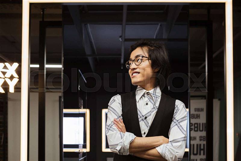 Photo of handsome young asian man wearing glasses dressed in jacket and shirt standing with arms crossed indoors and looking aside. Coworking concept, stock photo