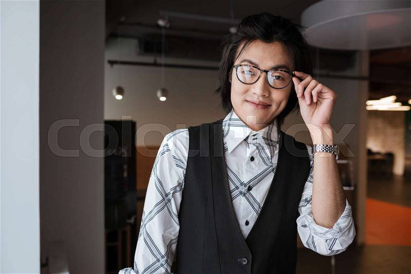 Image of happy young asian man wearing glasses dressed in jacket and shirt standing indoors and looking at camera. Coworking concept, stock photo