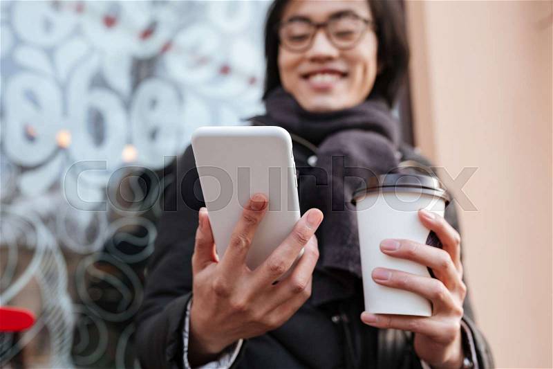 Picture of amazing young asian man wearing glasses and scarf walking outdoors and looking at phone while drinking coffee. Focus on phone, stock photo