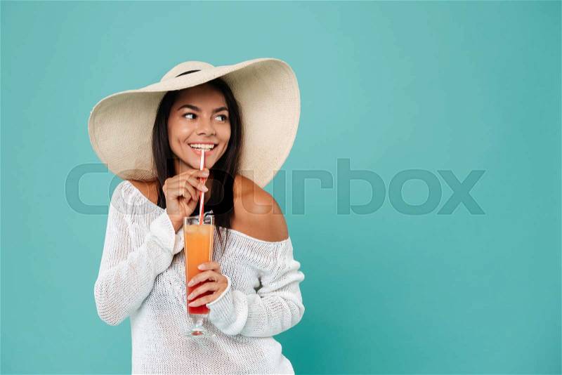 Cheerful beautiful young woman in hat drinking cocktail with straw over blue background, stock photo