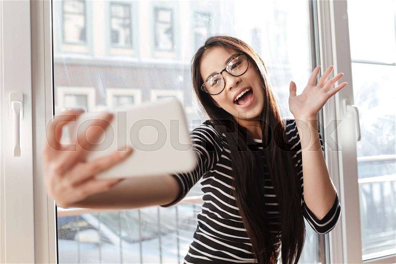 Funny Asian woman in eyeglasses which making selfie on phone, sitting on windowsill in cafeteria and waving, stock photo
