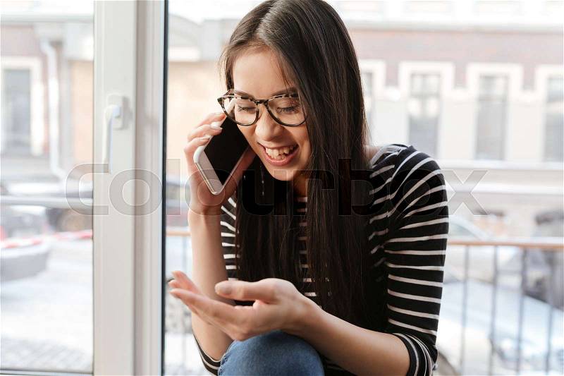 Happy Asian woman in eyeglasses which talking on phone and sitting on windowsill in cafeteria, stock photo