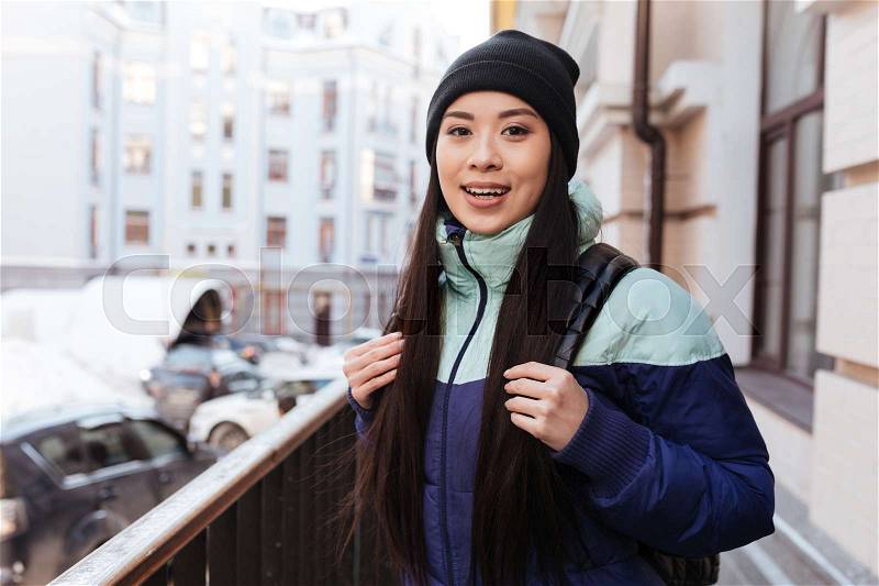 Happy Asian woman in warm clothes and with backpack which standing on the street and looking at camera, stock photo