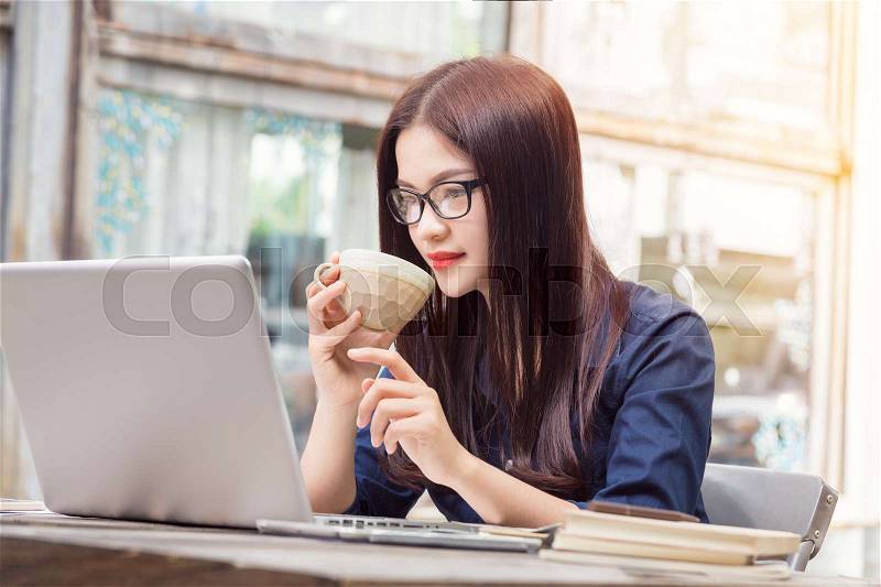 Young asian woman with eyeglasses using technology, looking at her laptop computer and holding cup of coffee, soft warm vintage tone, stock photo