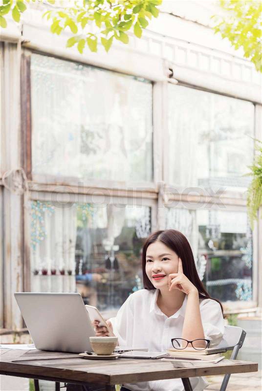Happy young asian woman thinking and enjoying her coffee while using her computer and smart phone, stock photo