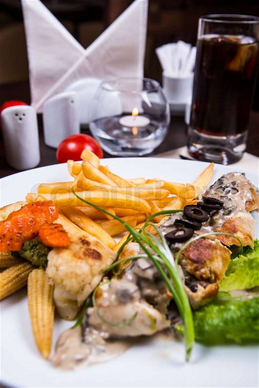 Chicken course with vegetables and french fries. Chicken dish, corn, broccoli, carrot, black olives, lettuce leaf. Salt, pepper, cola, serviette and candle on background, stock photo