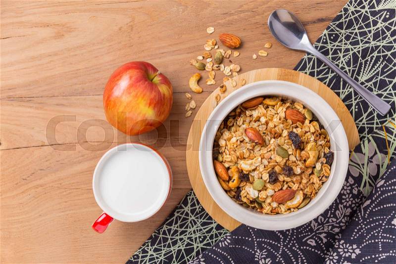 Bowl of cereal, cup of milk, and an apple on wooden table with spilled cereal, cloth napkin and silver spoon, room for text, stock photo
