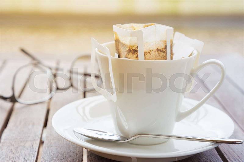 White coffee cup and spoon with white pre-packaged blended coffee in paper filter set on wooded table with out of focus blurred eyeglasses in background, stock photo