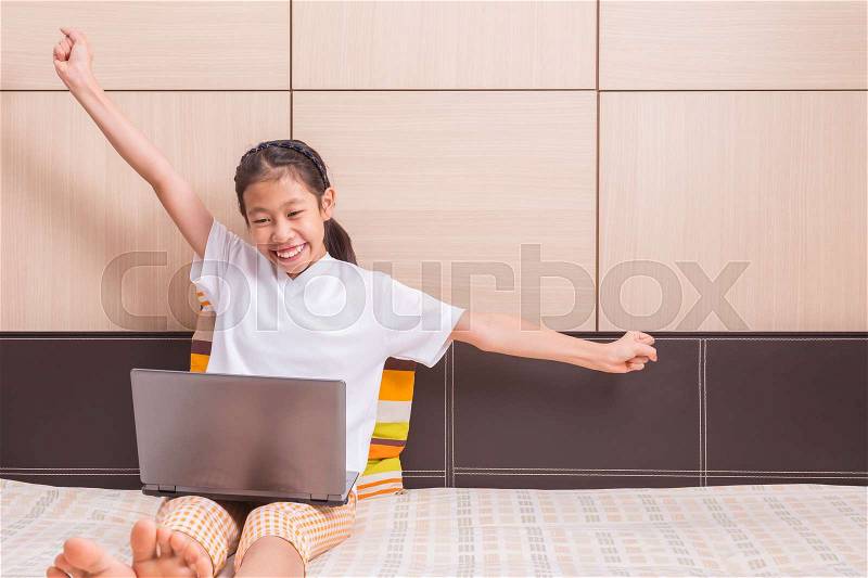 Happy smiling and excited asian girl using notebook computer to study on her bed, reaching out her arms in excited and successful pose, room for copy space text, stock photo